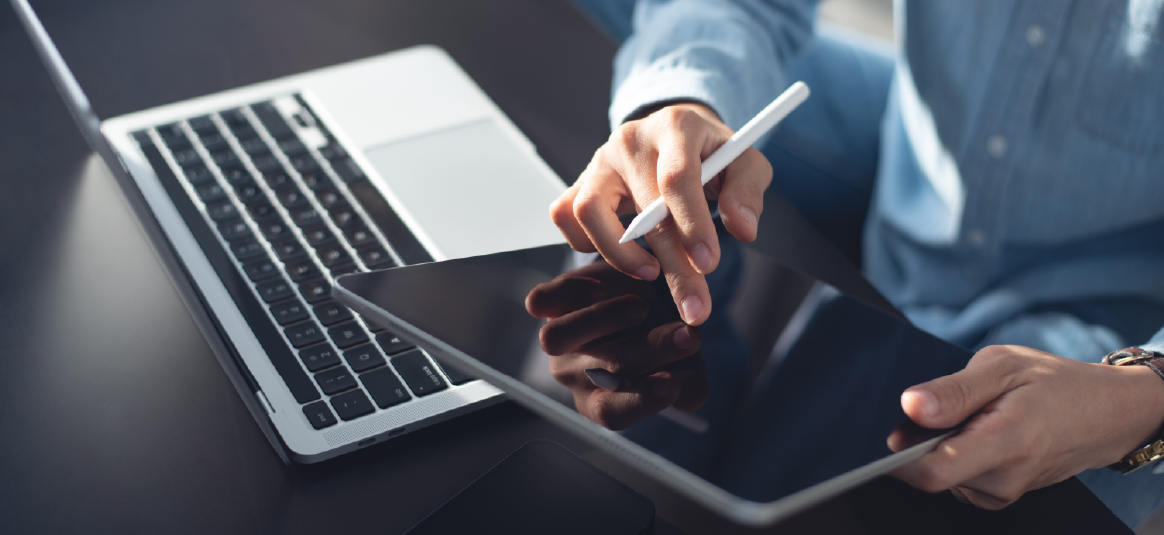 Person reading a touchscreen device and holding a stylus with a laptop in the background